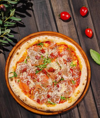 Tahtaya konmuş lezzetli bir pizza domates sosu, prosciutto ve peynir içerir. Etrafında taze fesleğen yaprakları ve kiraz domatesleri var. Canlı bir mutfak sunumu yapıyorlar..