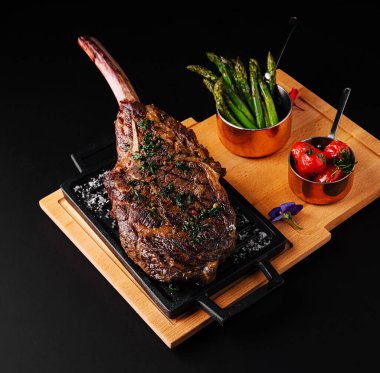 Mükemmelce pişirilmiş büyük bir baltalı biftek cızırdayan bir tabakta otlarla süslenir. Yanında ızgara kuşkonmaz ve kiraz domatesleri var. Gurme deneyimini geliştiriyor..