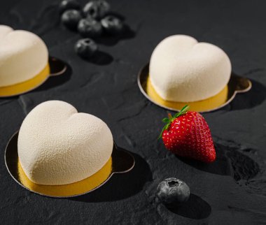 Kalp şeklindeki tatlılar zarif bir şekilde koyu bir tahta yüzeyde düzenlenir. Çevrelerinde taze yaban mersini ve canlı kırmızı bir çilek bulunmakta ve bu da hoş bir görsel karşıtlık yaratmaktadır..