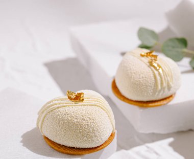 İki zarif beyaz tatlı desenli bir yüzeyde oturuyor, altın madalyaları ve pürüzsüz bir kaplaması var. Yumuşak ışıklandırma, lüks görünüşlerini geliştirir. İnce bir atmosfer yaratır..