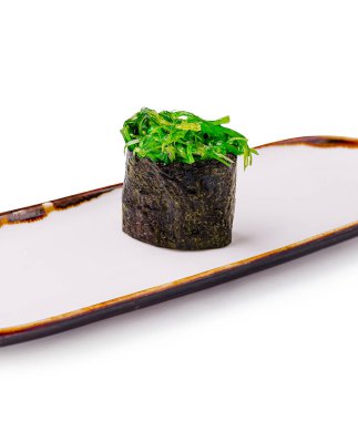 İyi sunulmuş bir suşi parçası, minimalist bir tabakta titiz bir mutfak sanatının sergilendiği canlı yeşil süslemelerle kaplanmış yosunlara sarılmış silindirik bir rulo sunar..