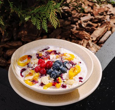 Yoğurt, çeşitli böğürtlenler ve taze mango dilimlerinden oluşan canlı bir meyve kasesi dışarıda, doğayla çevrili bir masada oturur. Güneş ışığı renkli sunumu güçlendirir.