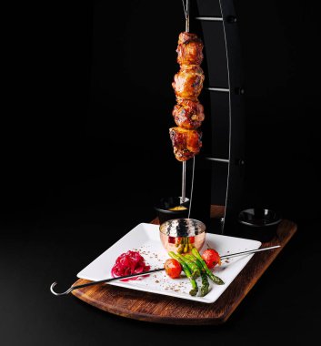 Lezzetli şişlenmiş et, canlı sebzeler ve soslarla dolu cilalanmış bir tabağın üzerinde asılı duruyor. Zarif sunum bu yemeğin lezzet ve mutfak sanatını vurguluyor.