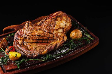 Mükemmel bir ızgara biftek, taze otlar ve kızarmış sebzelerle çevrili ahşap bir tepside duruyor. Tabakta, her et sever için ideal zengin ve lezzetli bir görünüm sergileniyor..