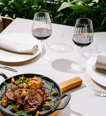 Izgara et ve sebzelerin yer aldığı bir gurme yemeği ahşap tabakta servis edilir. Yeşillik zemine kurulmuş yemeğe zarif kırmızı şarap kadehleri eşlik ediyor..