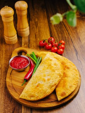 İki altın-kahverengi calzone, bir tabak domates sosu, taze biber, soğan ve kiraz domatesinin yanında ahşap bir kesim tahtasının üzerinde sunuluyor ve lezzetli bir yemek yaratıyor..