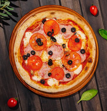 Taze domatesli, siyah zeytinli ve ahşap tahtada mozzarella soslu lezzetli bir pizza. Pizzayı çevreleyen kiraz domatesleri ve fesleğen yaprakları tazeliğini ön plana çıkarıyor..