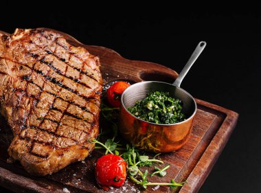 Mükemmel ızgara bir biftek ahşap bir tahtanın üzerinde oturur, yanında küçük bir kase canlı yeşil bitki sosu ve vişne domatesleri vardır. Sunum lezzetli yemeği vurguluyor..