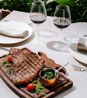Güzel bir şekilde sunulmuş ızgara biftek taze otların yanında ahşap bir tepside oturur. Yeşilliklerle çevrili şık bir açık hava yemek masasında iki kadeh kırmızı şarap bekliyor..