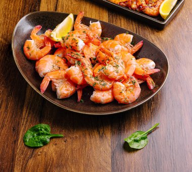 Bir tabak taze pişmiş karides güzel bir şekilde düzenlenir, otlarla süslenir ve ahşap bir masada limon dilimleriyle servis edilir. Yeşil yapraklar ayarlara renk katar.