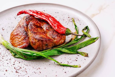 Taze yeşil soğan ve canlı kırmızı biberle süslenmiş güzel bir ızgara biftek gri bir tabakta oturur. Bu yemek, sağlıklı bir yemek için mükemmel olan zengin dokuları ve tatları ön plana çıkarıyor..