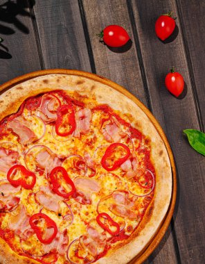 Jambonlu, kırmızı dolma biberli ve peynirli yeni pişmiş bir pizza, ahşap bir masada oturur. Kiraz domatesleri ve yeşil yapraklar ortama canlı renkler katıyor..