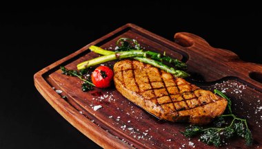 Mükemmel ızgara bir biftek ahşap bir tabağa konur ve yanında taze kuşkonmaz ve vişneli domates bulunur. Tuz ile tatlandırılmış bu yemek, zengin bir aroma ve çekici bir sunum yayar..