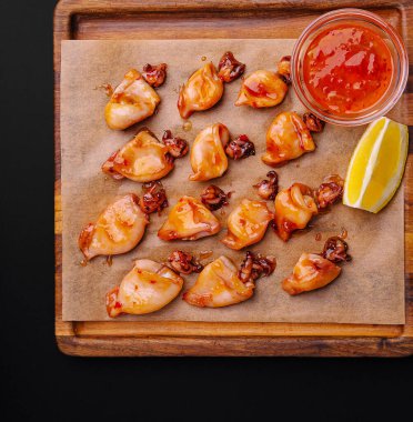 Ahşap tabakta tatlı ve baharatlı sos ve taze limon dilimleriyle süslenmiş ızgara kalamar parçaları sergileniyor. Tabakta canlı renkler ve nefis tatlar sergileniyor..