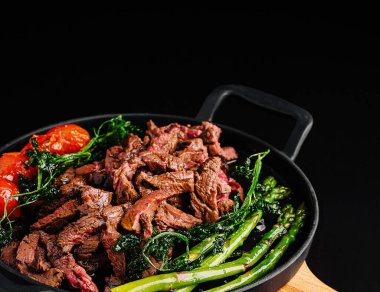 Izgara bifteğin yanında canlı kuşkonmaz ve tavada kızartılmış vişneli domates servis edilir. Tabakta taze malzemeler ve zengin renkler sergileniyor. Her yemek için mükemmel..