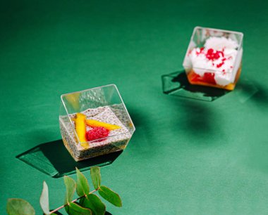 Renkli tatlılar yeşil bir masada berrak bardaklarda sergilenir. Bir fincan mango ve çilek içerirken, diğeri krem şanti ve kırmızı serpintilerle kaplı..