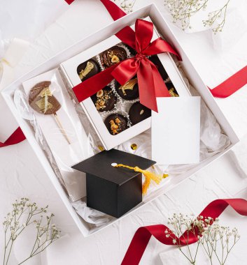 Mezuniyet kutlaması için içi gurme çikolataları, mezuniyet şapkası ve boş bir kartla dolu güzel düzenlenmiş bir hediye kutusu. Kırmızı kurdeleler ve dekoratif aksanlarla süslenmiş..