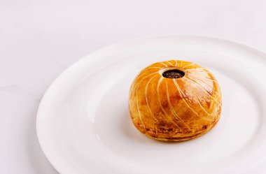 Yeni pişmiş bir hamur işi, altın-kahverengi kabuğunu ve karmaşık tasarımını sergileyen pürüzsüz beyaz bir tabakta tek başına oturur. Bu tatlı lezzetli bir deneyim vaat ediyor..