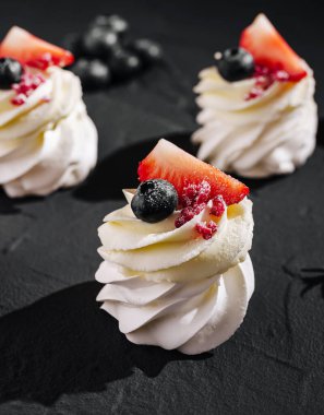 Çilek ve yaban mersiniyle süslenmiş çırpılmış kremalı şekerlemeler koyu bir yüzeye oturmuş, kremalı dokularını ve çekici bir ikram için canlı malzemelerini sergiliyorlar..