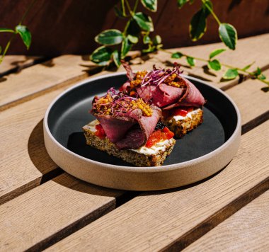 Yuvarlak tabakta iki tane açık yüzlü sandviç servis edilir. Kavrulmuş sebzeler ve otlar gibi canlı malzemeler sunuyorlar, kırsal bir ahşap zeminde güneş ışığına karşı dizilmiş..