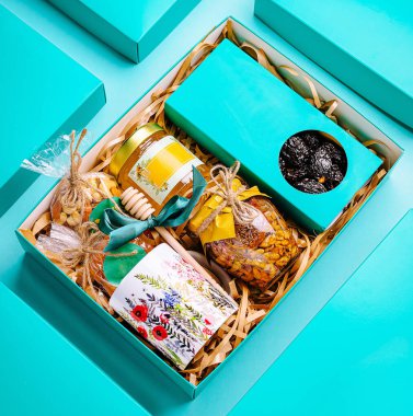 Özenle tasarlanmış bir hediye kutusunda bal kavanozları, kurutulmuş meyveler ve fındıklar var. Hepsi de güzel dekoratif aksanlarla sunulmuş. Canlı turkuaz ambalajlar davetkar bir görünüm yaratıyor..