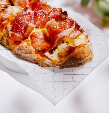 Erimiş peynirle kaplı kıtır kıtır bir pizza, tuzlu et ve beyaz bir tabakta bir tutam baharat var. Dekor asgari ve davetkar..