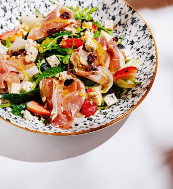 Canlı bir salata taze çilek, prosciutto ve parçalanmış peynirle karışık yeşilliklere sahiptir. Yemek zarif bir şekilde dekoratif bir kasede sunuluyor. Renkli malzemelerini vurguluyor..