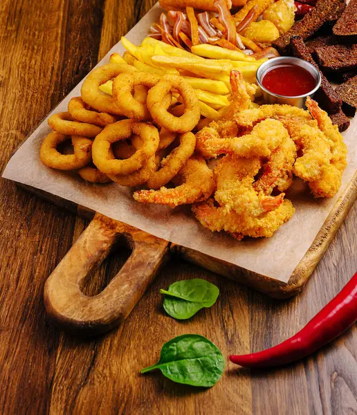 Lezzetli bir karides çeşidi, soğan halkaları ve ahşap bir tabakta dizilmiş altın patates kızartması. Tabak taze yeşillikler ve keskin bir sosla süslenmiş. Paylaşım için mükemmel..