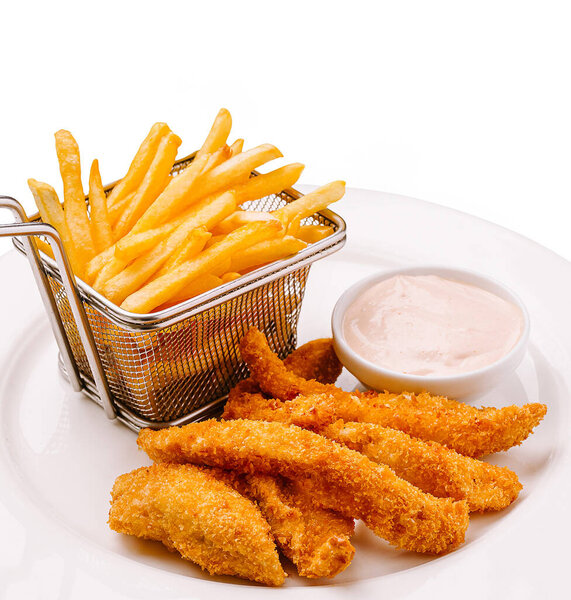 Crispy chicken tenders are presented alongside golden French fries in a small basket, accompanied by a dipping sauce. The meal is served in a casual dining setting.