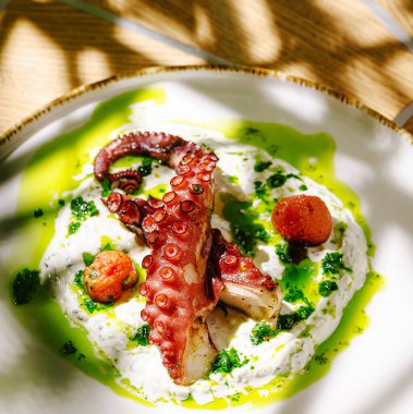 Yumuşak ızgara ahtapot, canlı yeşil otlar ve renkli domateslerle süslenmiş kremalı sosla güzelce düzenlenmiştir. Yemekler, aşçılık sanatını ve tazeliği gözler önüne seriyor..