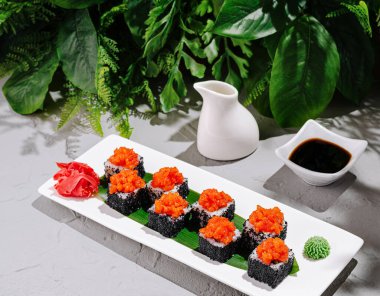 Büyüleyici bir suşi tabağında balık yumurtası ve wasabi ile yemyeşil bir zemin üzerine kurulmuş canlı suşi parçaları bulunuyor. Soya sosu ve küçük bir sürahi bu harika sunumu güçlendirir..