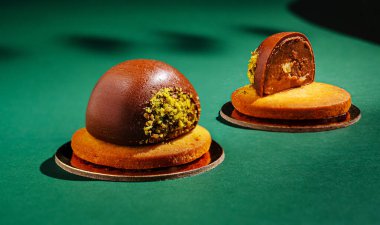 Uzmanca dizayn edilmiş iki tatlı, tatlı bir tabanın tepesinde fıstıklı parlak bir çikolata kubbesi barındırır. Parlak yeşil arkaplan çarpıcı renkleri ve karmaşık sunumu güçlendirir.