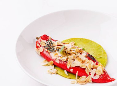 Dilimlenmiş badem ve siyah susam tohumlarıyla kaplı yeşil bir tortilla üzerinde canlı kırmızı biber servis edilir, dokular ve renklerin karışımı sunulur, gurme deneyimi için mükemmeldir..