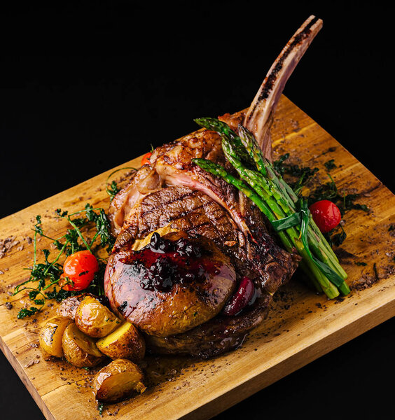 A perfectly cooked ribeye steak rests on a wooden board, accompanied by roasted potatoes, asparagus, and cherry tomatoes, garnished with herbs for a gourmet experience.