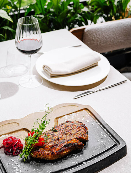 A beautifully plated grilled steak rests on a black stone, garnished with fresh herbs and tomatoes. A glass of red wine and an elegant white plate complete the inviting table setting.