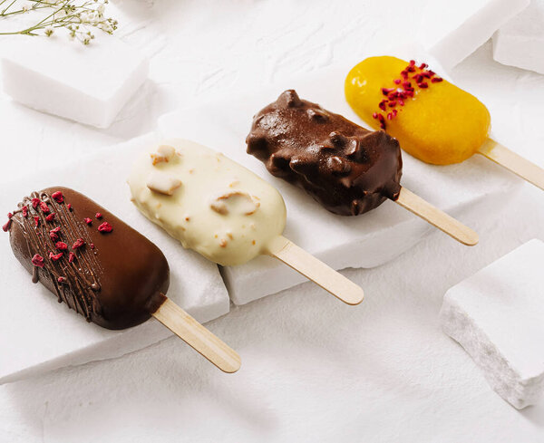 Четыре заманчивых леденца на деревянных палочках расположены на поверхности белого мрамора. Каждый из них имеет уникальное шоколадное покрытие, демонстрируя ароматы, такие как миндаль, темный шоколад, и манго.