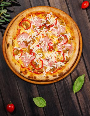 Yeni pişmiş bir pizza, üzerinde jambon, biber ve dilimlenmiş peynirle kahverengi ahşap bir masada oturur..