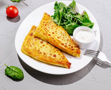 Taze yeşil salata ve küçük bir fincan kremalı sosun yanında iki altın sarısı doldurulmuş krep yer alır. Bir kiraz domates ve ıspanak yaprakları yemeğe canlı renk katar..