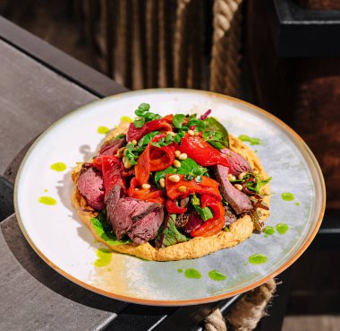 Bir tabakta kızarmış et, canlı kırmızı biber, taze otlar ve çam fıstığı dilimleri bulunan renkli bir yemek. Yemek, kırsal bir yemek düzenini ön plana çıkarıyor..