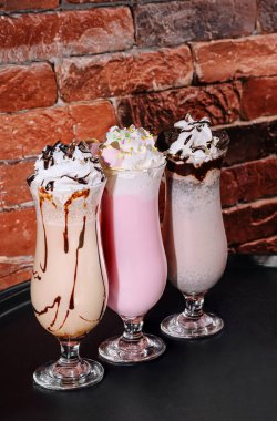 Üç renkli milkshake zarif bardaklarda servis edilir, her biri krem şanti ile kaplanır ve çikolatayla ıslanır. Kırsal bir tuğla duvara karşı sunuluyorlar, rahat bir ortam yaratıyorlar..