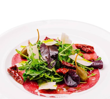 İnce dilimlenmiş dana carpaccio salatası, roka, güneşte kurutulmuş domates, zeytin ve tıraş edilmiş peynir, renkli ve iştah açıcı bir yemek yaratıyor..