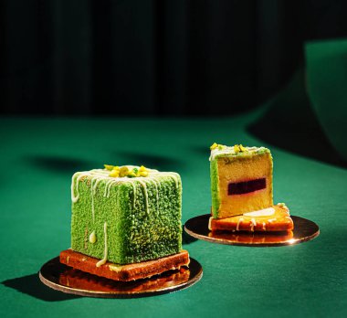 İki canlı yeşil tatlı karesi yansıtıcı tabaklarda zarif bir şekilde otururlar. Bir pasta çiselemeli, sergilenen katmanlar ve renkler ile süslenir. Kompozisyon zengin dokuları vurgular.