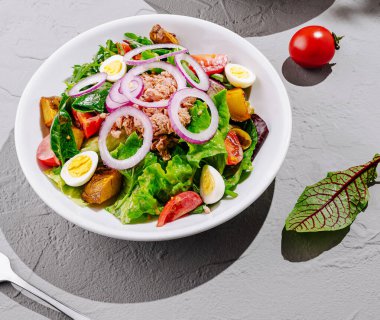 Renkli bir salata kasesinde taze yeşiller, kiraz domatesleri, dilimlenmiş turplar, haşlanmış yumurtalar ve dokulu bir yüzeyde ustalıkla sıralanmış ton balıkları yer alır. Yemekler sağlıklı beslenmeyi ön plana çıkarıyor..