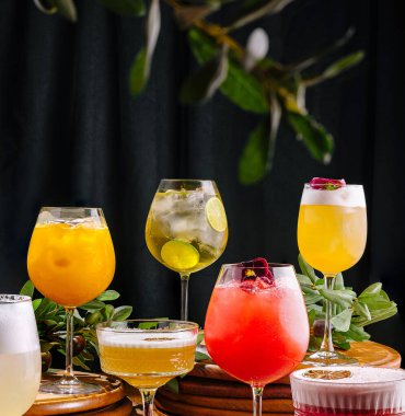 Çoklu canlı kokteyller yemyeşil ağaçlarla çevrili ahşap bir masada otururlar. İçkiler çeşitli renkler ve süslemeler sergiliyor, kutlama için uygun bir şenlik ortamı yaratıyor..