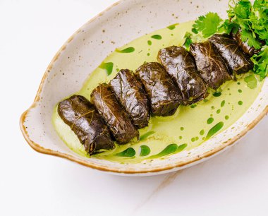 Çok güzel sarılmış doldurulmuş üzüm yaprakları, canlı yeşil sosla serpilmiş ve taze otlarla süslenmiş bir tabakta. Bu geleneksel yemek zengin lezzetler ve dokular sunar..