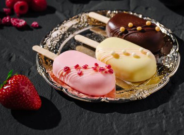 Üç zanaatkar dondurma gümüş bir tabakta, canlı renkler ve malzemeler sergiliyor. Tabağın etrafına taze çilek ve ahududular konur, görsel cazibeyi arttırır..