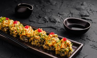 Suşi dürümleri siyah bir tabakta sanatsal olarak düzenlenir, üzerine canlı baharatlı mayonez ve kırmızı balık yumurtası serpiştirilir, yanında da desenli bir yüzey üzerindeki küçük tabaklardaki soya sosu ile birlikte..