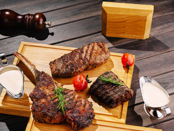 A selection of perfectly grilled meats presented on a wooden board with fresh herbs and roasted tomatoes. Accompanied by a creamy sauce, this dish invites indulgence and enjoyment.