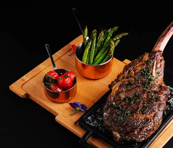 Izgara domates ve kuşkonmazın yanında güzel bir kaplanmış tomahawk bifteği sunulur. Tabak otlarla süslenmiş, marifetli mutfak sanatları ve canlı renkler sergileniyor..