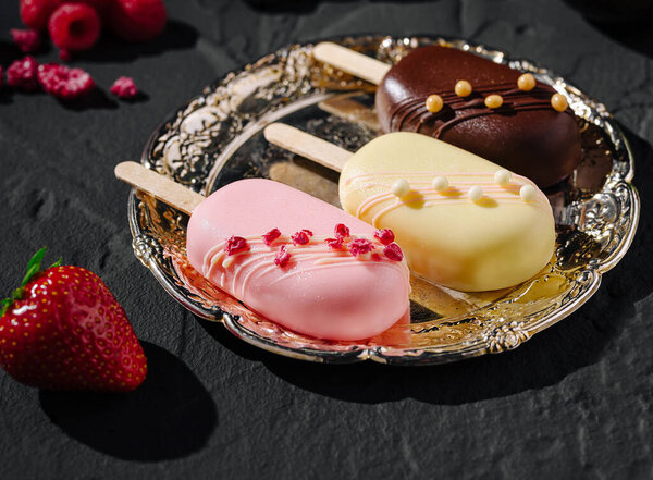 Три ремесленных мороженых сидят на серебряной тарелке, демонстрируя яркие цвета и начинки. Свежая клубника и малина размещены вокруг плиты, повышая визуальную привлекательность.